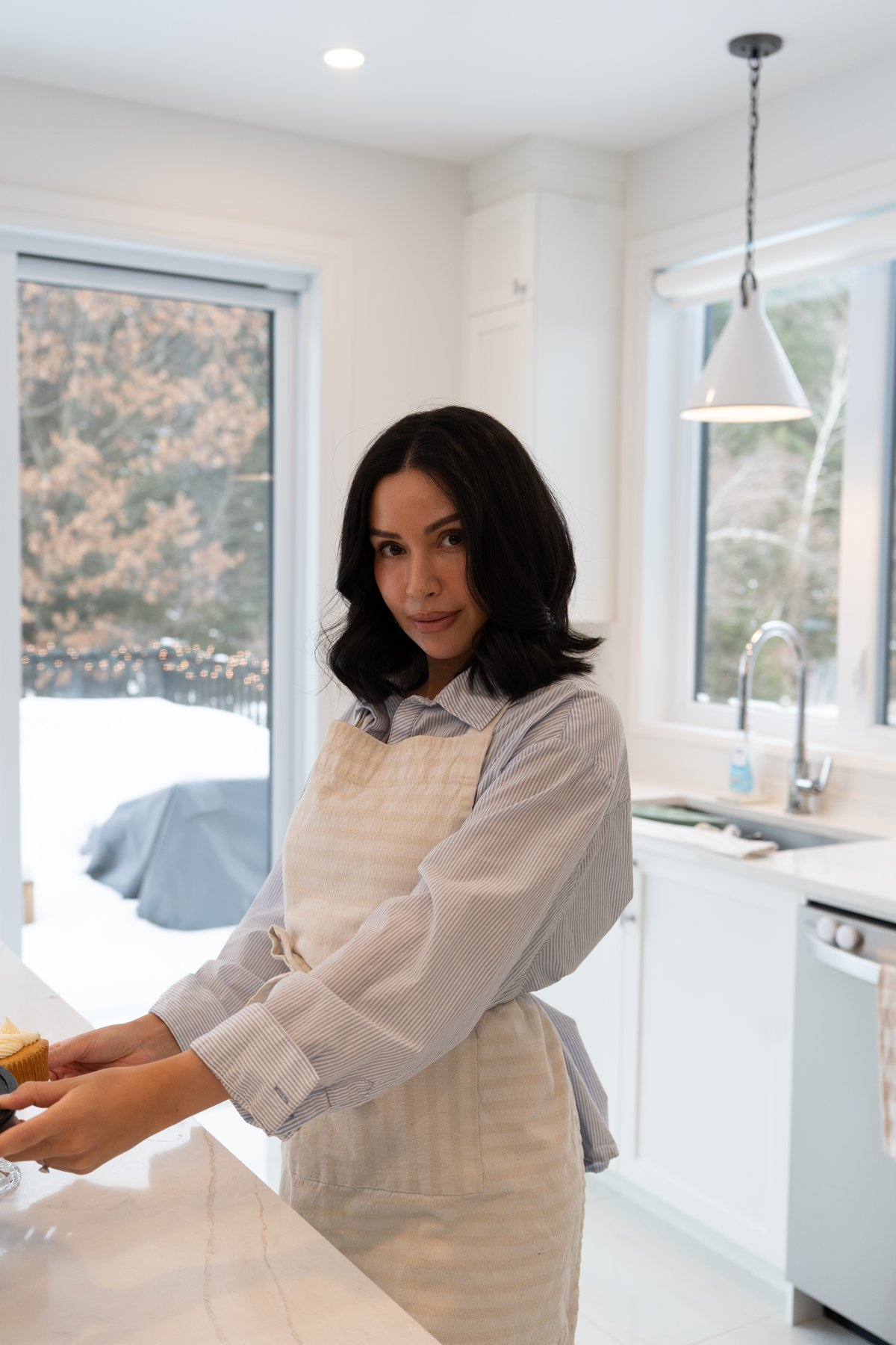 Kiana-Marie, founder of Oak & Rose Home, at home in a sunlit kitchen wearing a cream linen apron over a soft blue striped shirt.